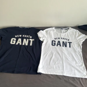 2-pack GANT t-shirts med tryck - Två snygga GANT t-shirts med 'NEW HAVEN GANT' tryck på bröstet. En är vit med marinblå text och en är mörkblå med vit text. Båda har rund halsringning med randig detalj på insidan och är i mjuk bomull. Perfekta för en clean och sportig look. Båda är i samma storlek 