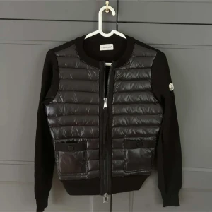 Moncler Cardigan  - Snygg svart pufferjacka från Moncler med quiltad front och ärmar i mjukt tyg. Jackan har två fickor med knappar, rund halsringning och dragkedja framtill. Perfekt för dig som vill ha en stilren och sportig look med exklusiv känsla.