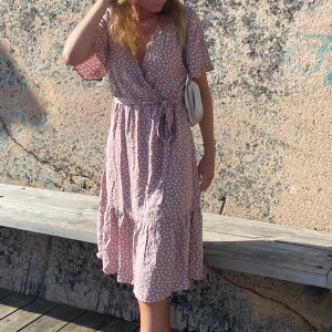 Ljusrosa prickig omlottklänning - Superfin ljusrosa omlottklänning med vita prickar och knyt i midjan. Klänningen har korta ärmar, v-ringning och volang nertill. Perfekt för varma dagar och ger en söt och avslappnad look.