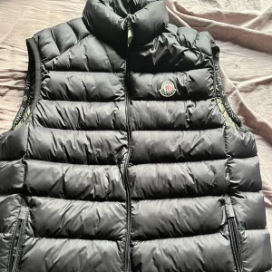 Svart dunväst från Moncler - Säljer en svart dunväst från Moncler med hög krage och dragkedja framtill. Västen har broderad Moncler-logga på bröstet och diskreta logoband längs ärmöppningarna. Insidan är olivgrön med en unik serietidningsetikett. Perfekt för lager-på-lager-stil. Frågar efter 3500 men kan pris kan diskuteras