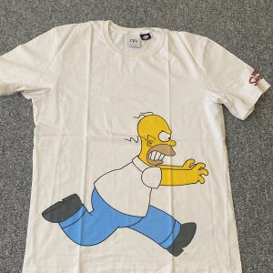 Vit Simpsons t-shirt från Zara - Vit t-shirt från Zara med stort tryck av Homer och Bart Simpson som jagar varandra, samt The Simpsons-logga på ärmen. Klassisk rund hals och korta ärmar. Perfekt för dig som älskar tecknade serier och vill sticka ut med en rolig printad tee.