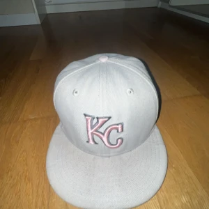 Beige KC keps från New Era - Snygg beige keps från New Era med broderad KC-logga i rosa och svart framtill. Kepsen har platt skärm, broderade ventilationshål och rosa detaljer på toppen och sidan. På sidan finns även ett broderat emblem med blommor. Perfekt för dig som gillar streetwear.