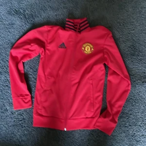 Manchester United jacka från Adidas - Röd Manchester United jacka från Adidas med klubbens broderade emblem på bröstet och svarta Adidas-loggan. Jackan har hel dragkedja, ståkrage med svarta ränder och två fickor framtill. Perfekt för dig som älskar fotboll och vill visa ditt lagstöd.