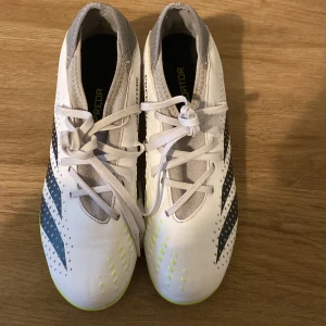 Adidas Predator vita fotbollsskor - Snygga Adidas Predator fotbollsskor i vitt med svarta och neongula detaljer. Skorna har snörning, låg profil och mönstrad ovandel för extra grepp. Perfekta för dig som vill sticka ut på planen med stilrena och sportiga skor.
