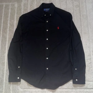 Svart skjorta från Ralph Lauren Slim Fit - Svart långärmad skjorta från Ralph Lauren i slim fit-modell. Klassisk design med knappar framtill och röd broderad logga på bröstet. Skjortan är tillverkad i bomull och har en stilren look som passar perfekt till jeans eller chinos.