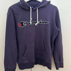 Lila Champion hoodie med broderad logga - Säljer en lila hoodie från Champion med stor broderad logga på bröstet. Tröjan har huva med vita snören och en klassisk känguruficka framtill. Perfekt för dig som gillar streetwear och vill ha en chill look.