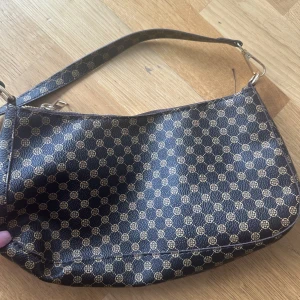 Brun  axelväska med mönster - Snygg brun axelväska med guldigt, cirkulärt mönster över hela väskan. Väskan har en smidig dragkedja upptill och ett klassiskt format axelband. Materialet är syntet och insidan är fodrad i svart tyg. Perfekt för dig som gillar stilrena detaljer.