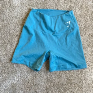Turkosa träningstights shorts från Gymshark - Säljer ett par turkosa träningstights shorts från Gymshark. De har hög midja och är tillverkade i ett stretchigt syntetmaterial som sitter tight. Perfekta för gymmet eller annan träning💕. De är uppsprättade längst ner men är inget man ser när de är på.