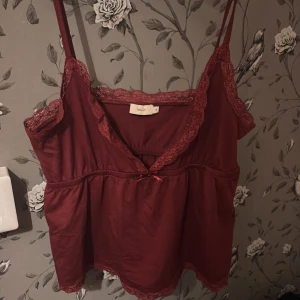 Vinröd linnetopp med spets från Nelly - Supersöt vinröd linnetopp från Nelly med tunna axelband och spetsdetaljer längs kanterna. Toppen har en liten rosett framtill och är lätt utställd nertill för en avslappnad look. Perfekt att styla med jeans eller kjol för en trendig vibe.