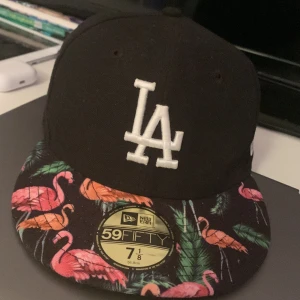 LA Dodgers keps New Era flamingo - Svart LA Dodgers 59FIFTY keps från New Era med broderad vit LA-logga framtill. Skärmen har ett färgstarkt flamingomönster med gröna blad och rosa-orange flamingos. Klassisk basebollform och broderade loggor på sidan och bak. Perfekt statement-piece för dig som gillar unika detaljer.