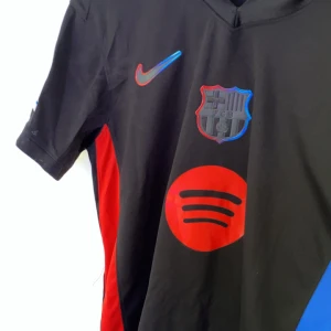 FC Barcelona Lamine Yamal tröja Nike - Säljer en svart FC Barcelona fotbollströja från Nike med Lamine Yamal och nummer 19 på ryggen. Tröjan har röda detaljer, Spotify-logga på bröstet och Champions League-märke på ärmen. Materialet är lätt och andas, perfekt för match eller träning.