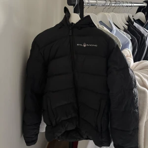 Svart pufferjacka från Sail Racing - Svart pufferjacka från Sail Racing med huva och logga på bröstet samt bak på huvan. Jackan har en klassisk quiltad look och dragkedja framtill. Perfekt för kalla dagar när du vill hålla stilen och värmen. S/M