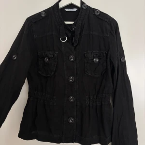 Svart linneskjorta från Marella Sport - Svart linneskjorta från Marella Sport med markerad midja, stora knappar och två bröstfickor med lock. Skjortan har långa ärmar med knappdetaljer och coola axelpartier. Perfekt för dig som gillar en stilren och edgy look.