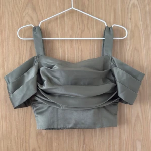 Grågrön satin topp från Bubbleroom - Fin grågrön croppad topp från Bubbleroom i satinliknande material. Toppen har breda axelband, draperad framsida, veckade detaljer på ärmarna och dragkedja baktill. Använd 1 gång. 