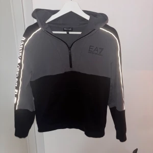 EA7 hoodie med reflexdetaljer - EA7 Emporio Armani hoodie i svart och grått med halv dragkedja och huva. Reflexdetaljer längs ärmarna och stort logotryck på ena ärmen. magficka framtill. 