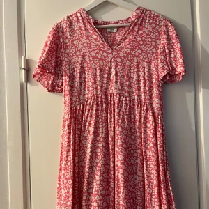 Rosa blommig kortklänning från JDY - Supersöt rosa och vit kortklänning från JDY med blommigt mönster. Klänningen har v-ringning, korta ärmar och en luftig passform som ger en avslappnad vibe. Perfekt för varma dagar när du vill känna dig både bekväm och stylish.