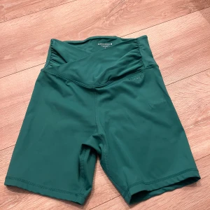 Gröna träningstights-shorts Björn Borg - Snygga gröna träningstights-shorts från Björn Borg med hög midja och omlott-detalj framtill. De är tillverkade i ett stretchigt syntetmaterial som sitter tight och bekvämt. Perfekta för gymmet eller löprundan!