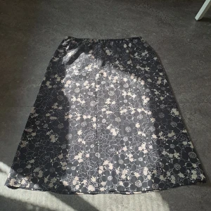 Svart blommig långkjol, vintage - Svart långkjol från Brandtex med beige och vita blommor i ett snyggt mönster. Kjolen har resår i midjan för en bekväm passform och är tillverkad i ett lätt och mjukt tyg. Perfekt för dig som gillar stilrena plagg. Materialet är väldigt svalt och det finns en innerkjol. 