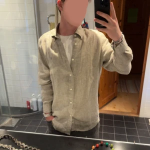 Beige linneskjorta med knappar - Snygg beige skjorta i linne med klassisk krage och vita knappar. Skjortan har lång ärm och en avslappnad passform, perfekt att bära över en t-shirt för en chill look. Enkel och stilren design som passar till många outfits.