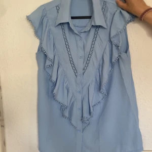 Ljusblå ärmlös blus med volang - Supersöt ljusblå blus utan ärmar med volangdetaljer längs framsidan och axlarna. Blusen har knappar framtill och dekorativa spetsinslag som ger en romantisk vibe. Perfekt för dig som gillar en feminin och trendig look. Endast testad