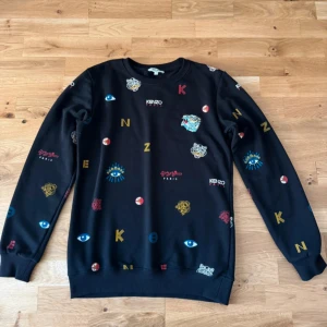 Svart Kenzo sweatshirt med broderier - Svart sweatshirt från Kenzo med färgglada broderade detaljer som ögon, bokstäver och tigrar. Tröjan har rund hals, långa ärmar och ribbade muddar. Tillverkad i 100% bomull för en skön och avslappnad känsla. Perfekt för dig som gillar statement-plagg.