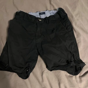 Svarta shorts från Gant - Svarta shorts från Gant med klassisk design och avslappnad passform. De har fickor fram och bak med knappdetaljer, samt uppvikta benslut. Tillverkade i mjuk bomull, perfekta för varma dagar. Snygga och stilrena shorts som funkar till det mesta.