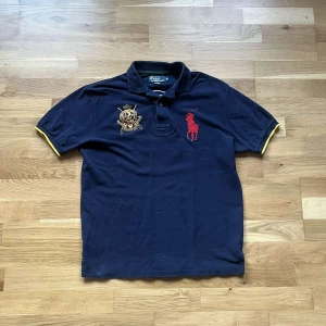 Ralph lauren piké - Snygg mörkblå pikétröja från Polo Ralph Lauren med röd broderad logga och gul detalj på ärmsluten. Stort vitt nummer 2 på ryggen och broderat emblem framtill. Klassisk krage och korta ärmar. Perfekt för dig som gillar sportig och preppy stil.
