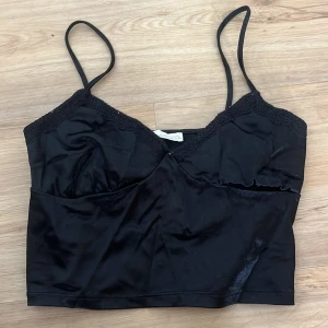 Svart linnetopp från Pull&Bear - Snygg svart linnetopp från Pull&Bear med tunna axelband och spetsdetaljer längs v-ringningen. Toppen har en glansig satinliknande yta och croppad passform, perfekt att styla med jeans eller kjol. Enkel men ändå elegant look. Så fint för att gå ut.