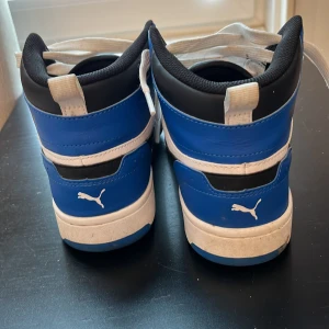 Puma sneakers i blått och svart, strl 39 - Snygga Puma sneakers i blått, svart och vitt med högt skaft och klassisk snörning. Skorna har SoftFoam+ innersula för extra komfort och cool streetstil. Perfekta för dig som gillar sportig vibe och vill sticka ut med färg. Puma-logga på sidan och hälen.