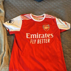 Arsenal Ødegaard 8 matchtröja - Röd och vit Arsenal fotbollströja från Adidas med Ødegaard och nummer 8 på ryggen. Klassisk design med vita ränder på axlarna, klubbmärke på bröstet och Premier League-märke på ärmen. Tillverkad i lätt och ventilerande material.