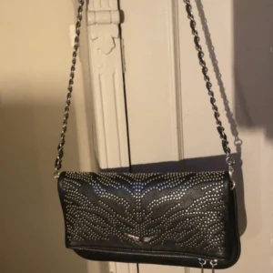 Zadig & Voltaire väska - Snygg svart axelväska från Zadig & Voltaire i skinn med silverfärgade nitar i coolt mönster på framsidan. Kedjerem i metall och dragkedja baktill. Perfekt storlek för mobilen och det viktigaste.