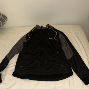 Svart och grå sportjacka från Kipsta - Sportig svart och grå vindjacka från Kipsta med gula detaljer. Jackan har dragkedja framtill, långa ärmar och två fickor. Perfekt för träning eller utomhusaktiviteter. Materialet är lätt och känns smidigt på kroppen.