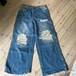 Wide ripped jeans från We Be Free - Blåa wide leg jeans från We Be Free med stora slitningar och trasiga detaljer på benen. Jeansen har hög midja, fem fickor och råa kanter nertill. Tillverkade i klassisk denim med coolt sliten look. Dem e väldigt rare och de säljs inte längre