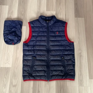 Mörkblå dunväst från Ralph Lauren - Passar mellan 160-175 S model mycket bra skick 9/10.  Snygg mörkblå dunväst från Ralph Lauren med röda detaljer och dragkedja framtill. Västen har två fickor med dragkedja och klassisk broderad logga på bröstet. Kommer med tillhörande påse för smidig förvaring. Perfekt för lager-på-lager-stil.
