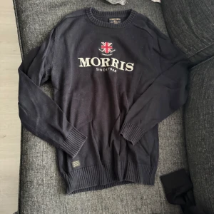 Mörkblå stickad tröja från Morris - Snygg mörkblå stickad tröja från Morris med broderad logga och brittisk flagga på bröstet. Tröjan har rund halsringning, långa ärmar och ribbade muddar vid ärmslut och nederkant. Perfekt för dig som gillar klassisk och stilren look.