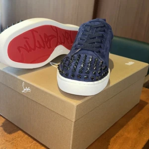 Blå Louboutin sneakers med nitar - Snygga blå sneakers från Louboutin i mocka med svarta nitar på tån och klassisk röd sula. Skorna har vita sulor, snörning och en riktigt lyxig vibe. Perfekta för dig som vill sticka ut med exklusiva detaljer och streetstyle-känsla.
