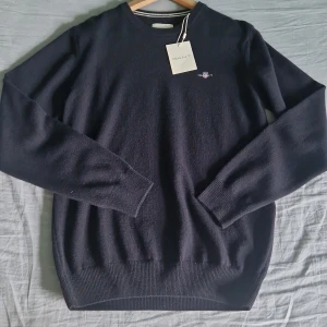 Svart lammullströja från GANT - Klassisk svart tröja från GANT i superfin lammull. Rund halsringning, diskret broderad logga på bröstet och ribbade muddar vid ärmslut och nederkant. Perfekt för dig som gillar stilrena och tidlösa plagg med premiumkänsla.