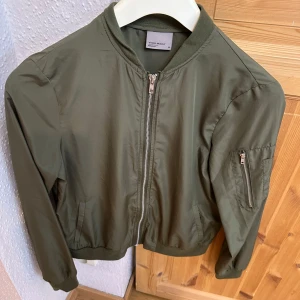 Olivgrön bomberjacka från Vero Moda - Snygg olivgrön bomberjacka från Vero Moda med klassisk passform. Jackan har dragkedja framtill, ribbade muddar och en ärmficka med dragkedja. 