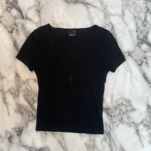 Svart ribbad topp från Gina Tricot - En svart, ribbad topp från Gina Tricot med korta ärmar och rundad halsringning. Toppen har tre knappar framtill som ger en snygg detalj.