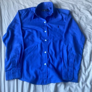Blå skjorta från Ralph Lauren - Klassisk blå skjorta från Ralph Lauren med lång ärm och knappar framtill. Skjortan har en snygg krage och ikoniska broderade loggan på bröstet. Tillverkad i mjuk bomull som känns skön mot huden. Perfekt för dig som gillar stilrena och tidlösa plagg.