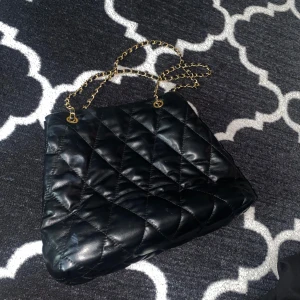 Svart quiltad axelväska med kedja - Säljer en svart quiltad axelväska i skinnimitation med guldfärgad kedjerem. Väskan har magnetlås och ljus insida med innerfack och dragkedja. Perfekt storlek för allt du behöver ha med dig och ger en lyxig vibe till din outfit.