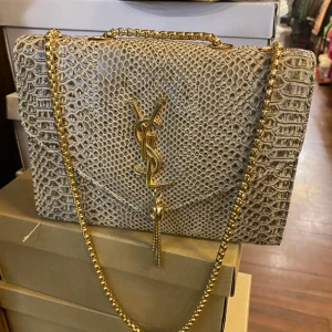 Beige axelväska i skinn - Säljer en beige axelväska  med lyxig ormskinnspräglad yta och guldfärgad kedja. Väskan har ikonisk loga  framtill och en snygg tofsdetalj. Perfekt storlek för det viktigaste och ger en exklusiv vibe till din outfit.