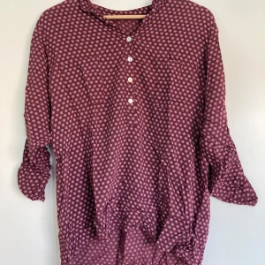 Mönstrad vinröd henley-skjorta - Vinröd henley-skjorta med små blommiga mönster i lila och rosa. Skjortan har knappar framtill, långärmade ärmar som kan rullas upp och fästas med knapp. Luftigt material och avslappnad passform, perfekt för en chill och snygg look.