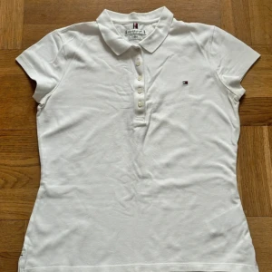 Vit pikéblus från Tommy Hilfiger - Klassisk vit pikéblus från Tommy Hilfiger med korta ärmar, krage och knappar framtill. Liten broderad logga på bröstet. Perfekt för en clean och sportig look.