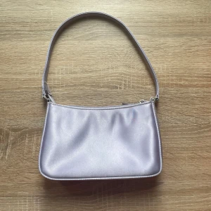 Ljuslila axelväska från Barratts - Snygg ljuslila axelväska från Barratts i satinliknande material. Väskan har en enkel, stilren design med dragkedja upptill och ett innerfack med dragkedja. Perfekt storlek för det viktigaste och en riktigt trendig accessoar.