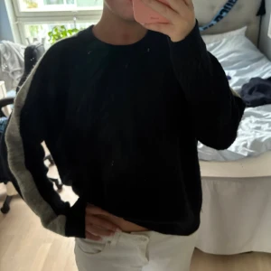 Blå stickad tröja med grå rand - Säljer en blå stickad tröja med en grå rand längs ärmarna. Tröjan har en avslappnad passform och är långärmad med rund hals.