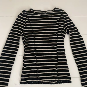 Randig långärmad topp från Gina Tricot - Svart och vit ribbad långärmad topp från Gina Tricot i storlek XXS. Tröjan har horisontella ränder, figurnära passform och vågiga kanter vid ärmslut och nederkant. Perfekt för dig som gillar en klassisk men trendig look.