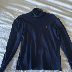 Marinblå polotröja från Polo Ralph Lauren - Snygg marinblå polotröja från Polo Ralph Lauren med klassisk broderad logga på bröstet. Tröjan är i mjuk bomull, har lång ärm och en hög polokrage. Perfekt för lager-på-lager och stilrena outfits.