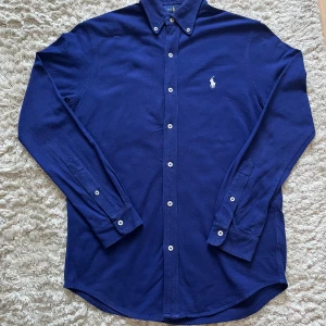 Ralph lauren skjorta - Klassisk marinblå skjorta från Ralph Lauren med vit broderad logga på bröstet. Skjortan har lång ärm, knappar framtill och button-down krage. Tillverkad i tunn bomull med featherweight mesh för extra komfort. Perfekt för att ha en stilren look nu i sommar.