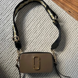 Marc Jacobs Snapshot axelväska beige - Snygg beige axelväska från Marc Jacobs med guldiga metalldetaljer och logga framtill. Väskan har en bred, justerbar axelrem i svart och guld med text, samt dragkedja upptill och praktiskt ytterfack. Väskan är endast provad. 